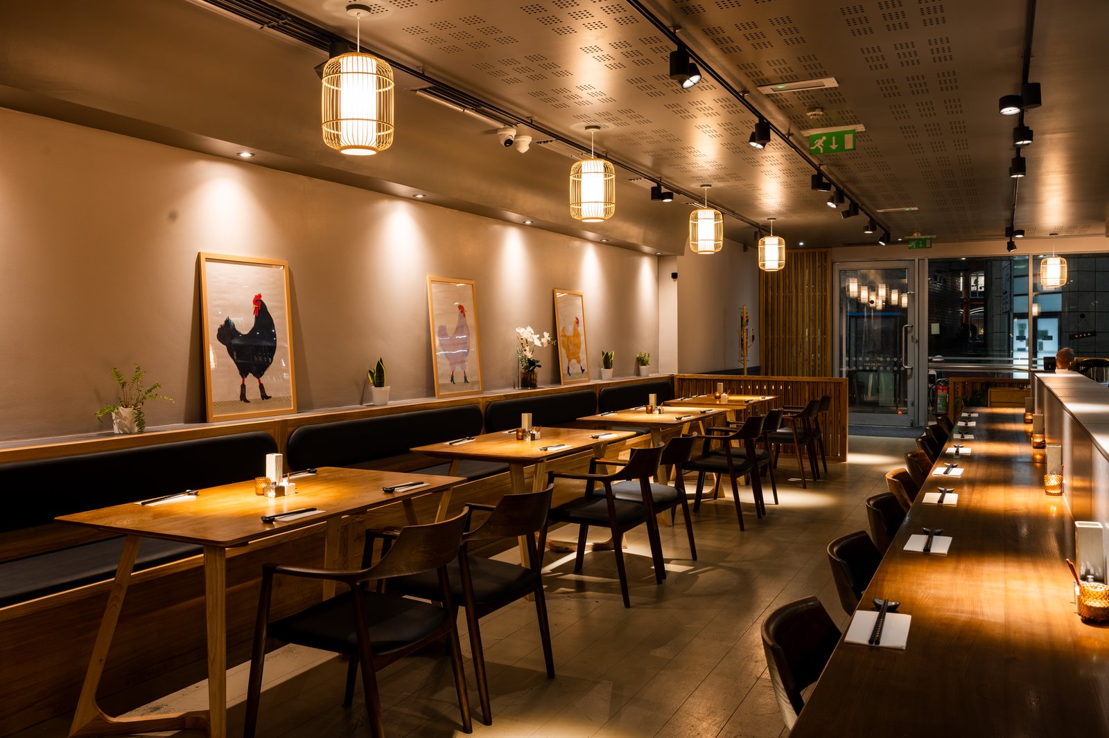 The Hotori dining room at dusk — warm pendant lights, banquette seating, and framed chicken paintings along the wall